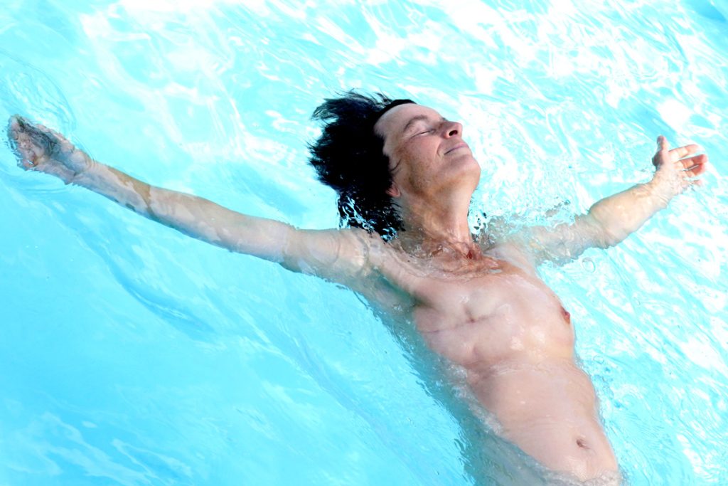 photo couleur une femme dans une piscine bras en croix faisant la planche visage apaisé les yeux fermés on voit son buste un sein manquant on voit nettement la cicatrice