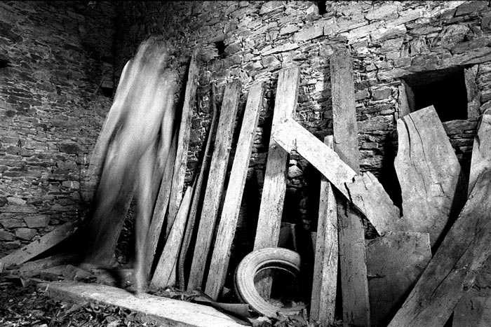 photo noir et blanc d'un vieux mur de pierres sur lequel reposent des planches et au sol un pneu laissant deviner la présence spectrale d'un corps humain se mêlant aux planches