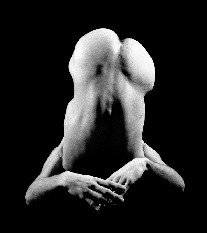 photo noir et blanc corps homme nu en position de demie chandelle mains croisées fesses en l'air donnant l'impression d'un masque ou sexe en érection